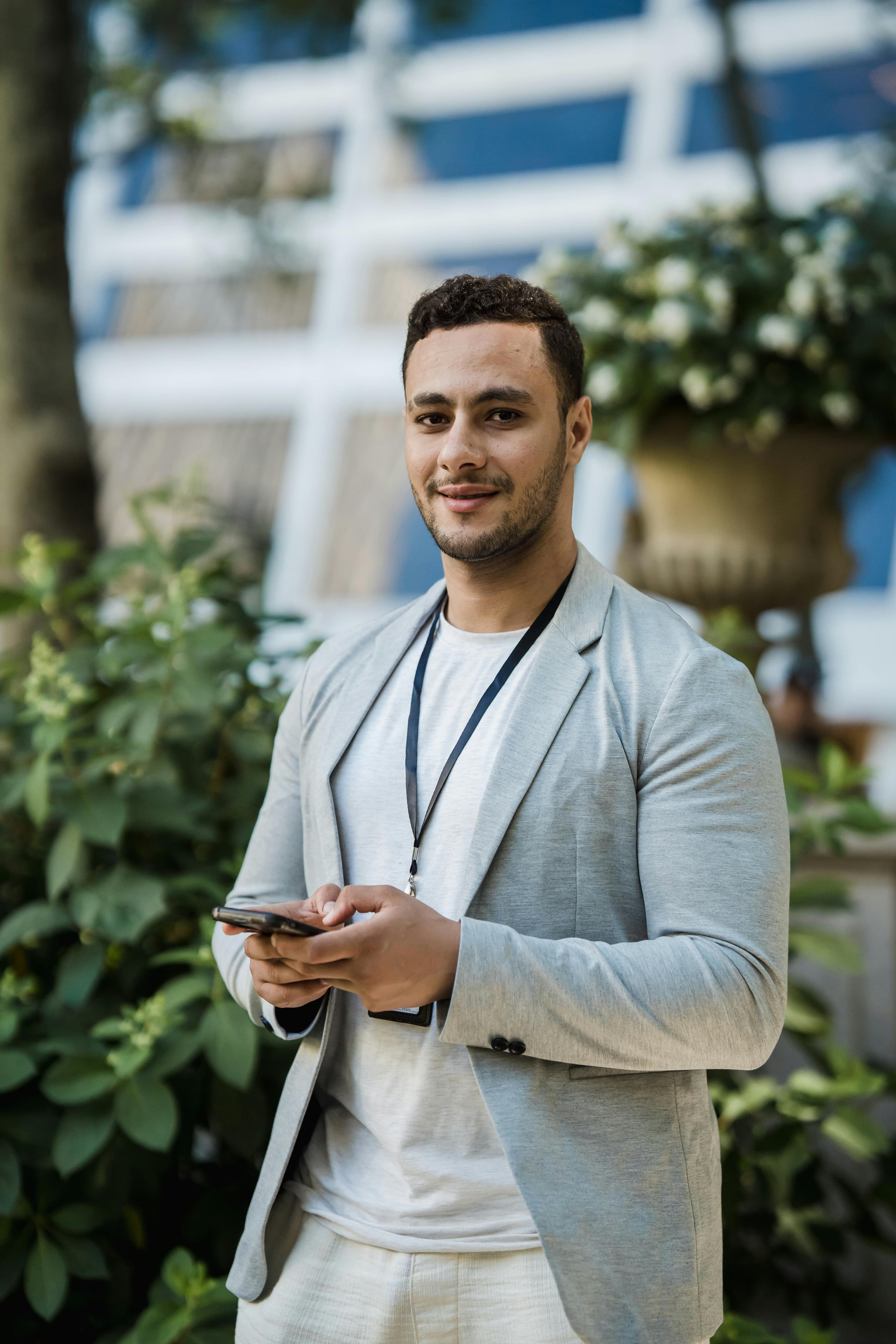 A young professional in a light blazer wearing a lanyard ID badge and holding a phone outdoors