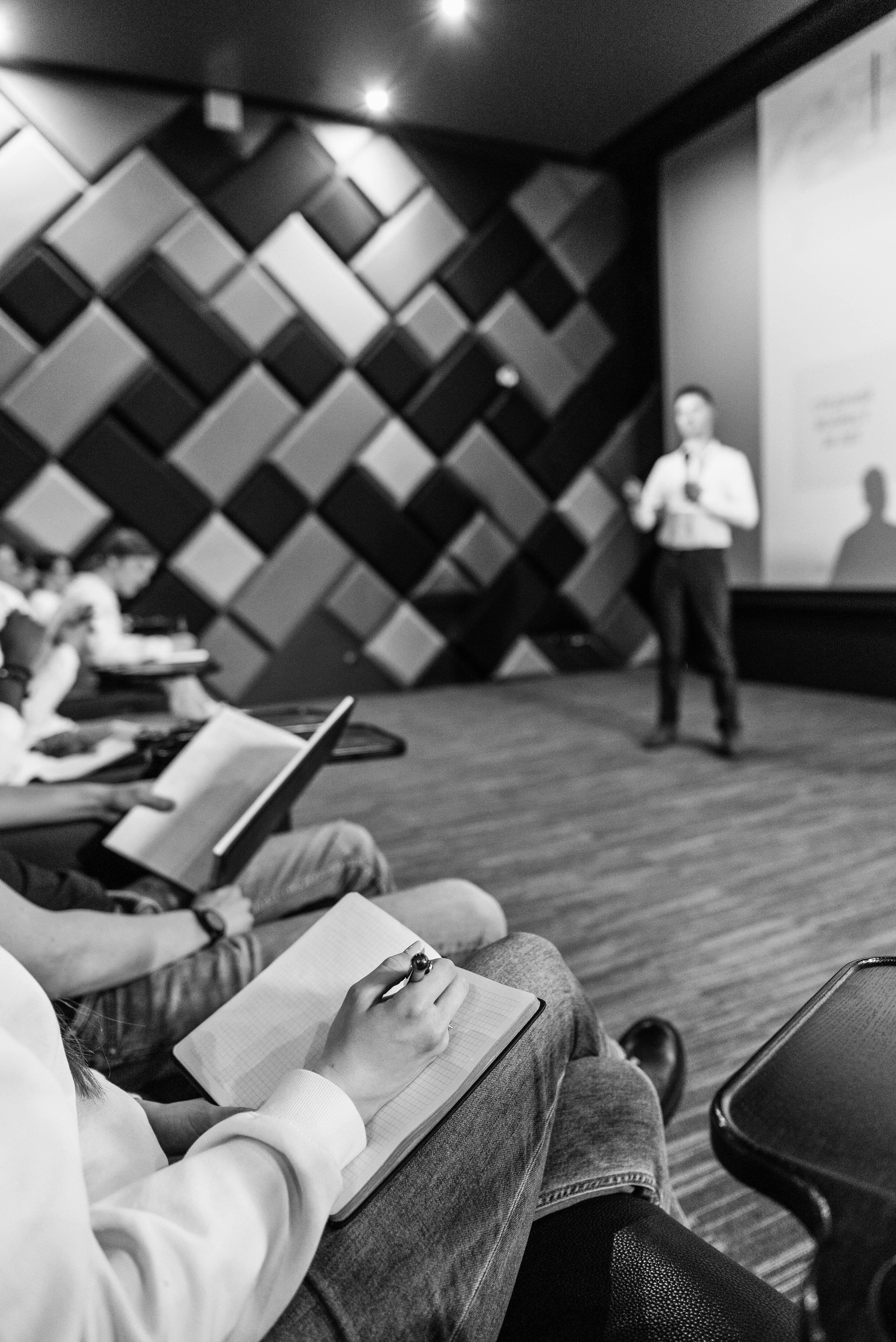 A speaker presenting at the front of a startup meetup while attendees take notes, in black and white