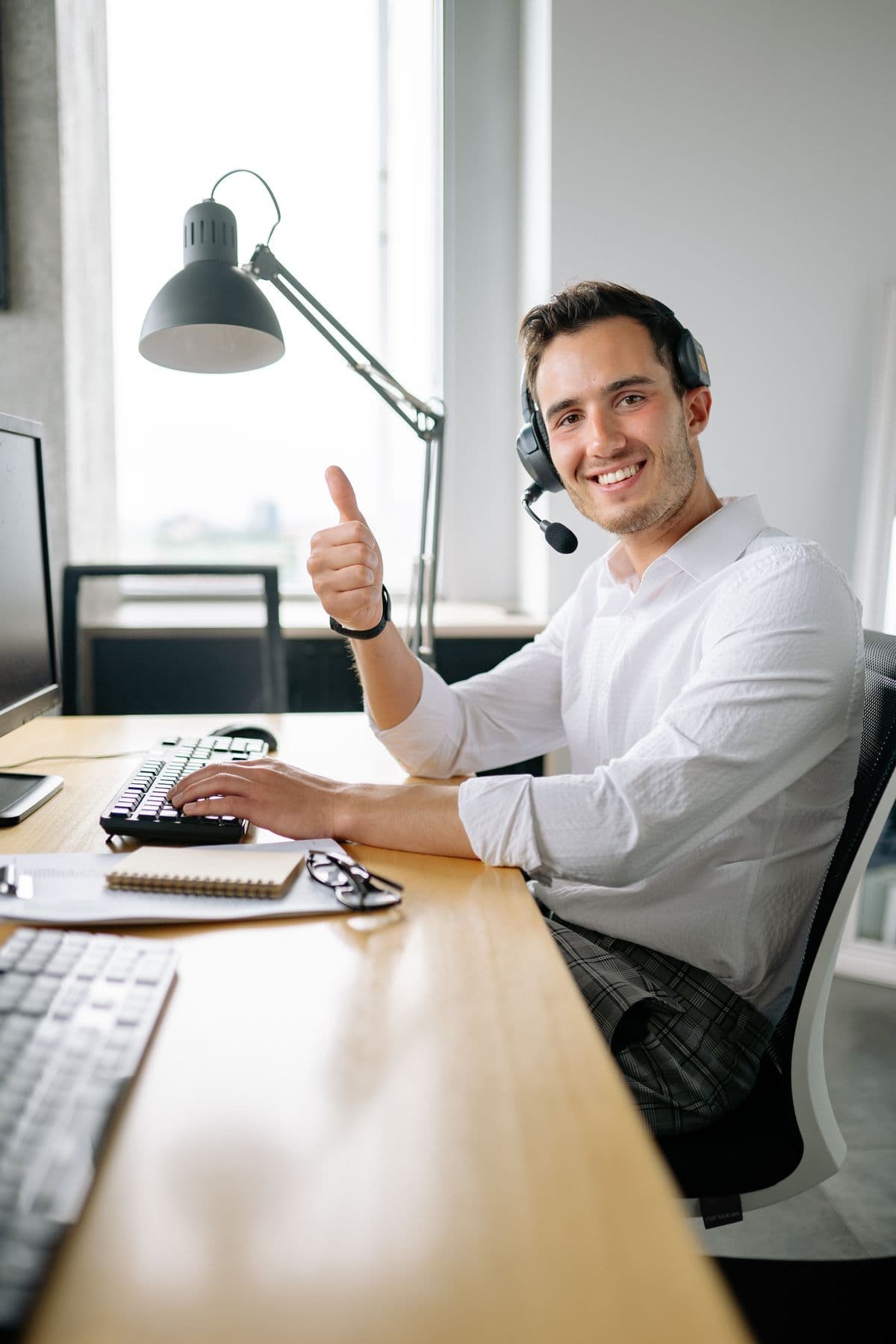 A smiling remote worker wearing a headset gives a thumbs up at a home-office desk
